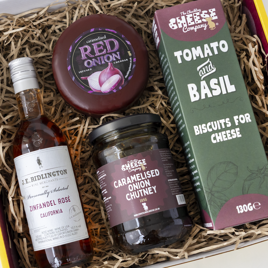 A curated cheese and snack gift box from The Chuckling Cheese Company, featuring a bottle of J.R., a jar of chutney, a red onion cheese truckle, and a box of biscuit for cheese.