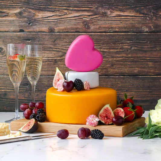 Chuckling Cheese celebration cheese cake tower with stacked cheese wheels, pink heart topper, berries, figs, flowers and champagne glasses