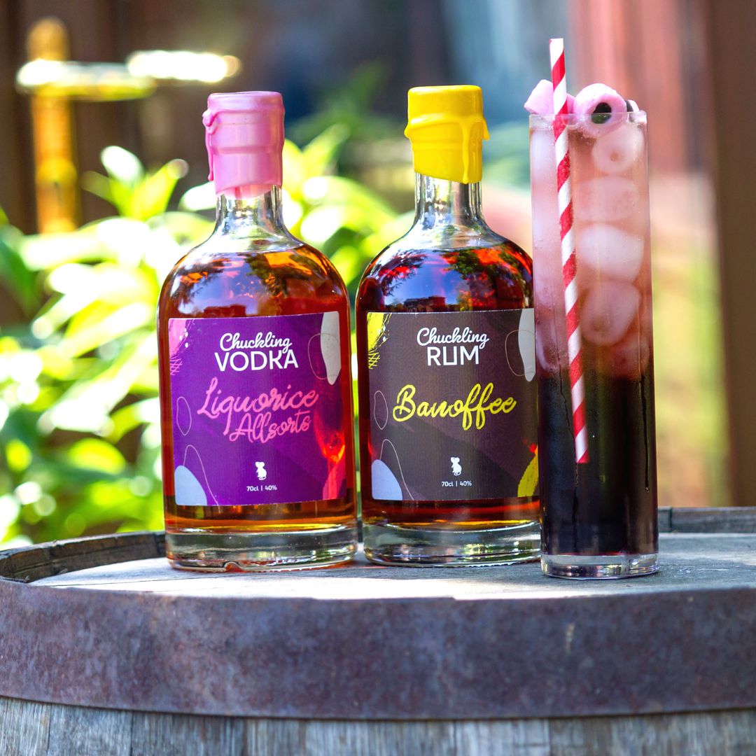 Liquorice Allsorts Vodka and Banoffee Rum bottles beside a tall layered cocktail on a wooden barrel