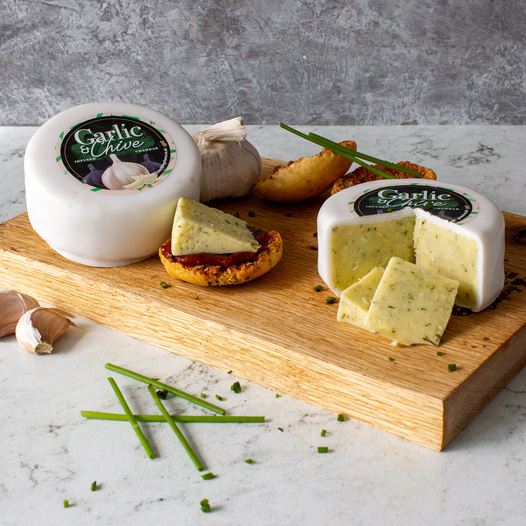 Chuckling Cheese Garlic & Chive Cheddar truckles with sliced cheese, garlic cloves, crackers and chives on a wooden board