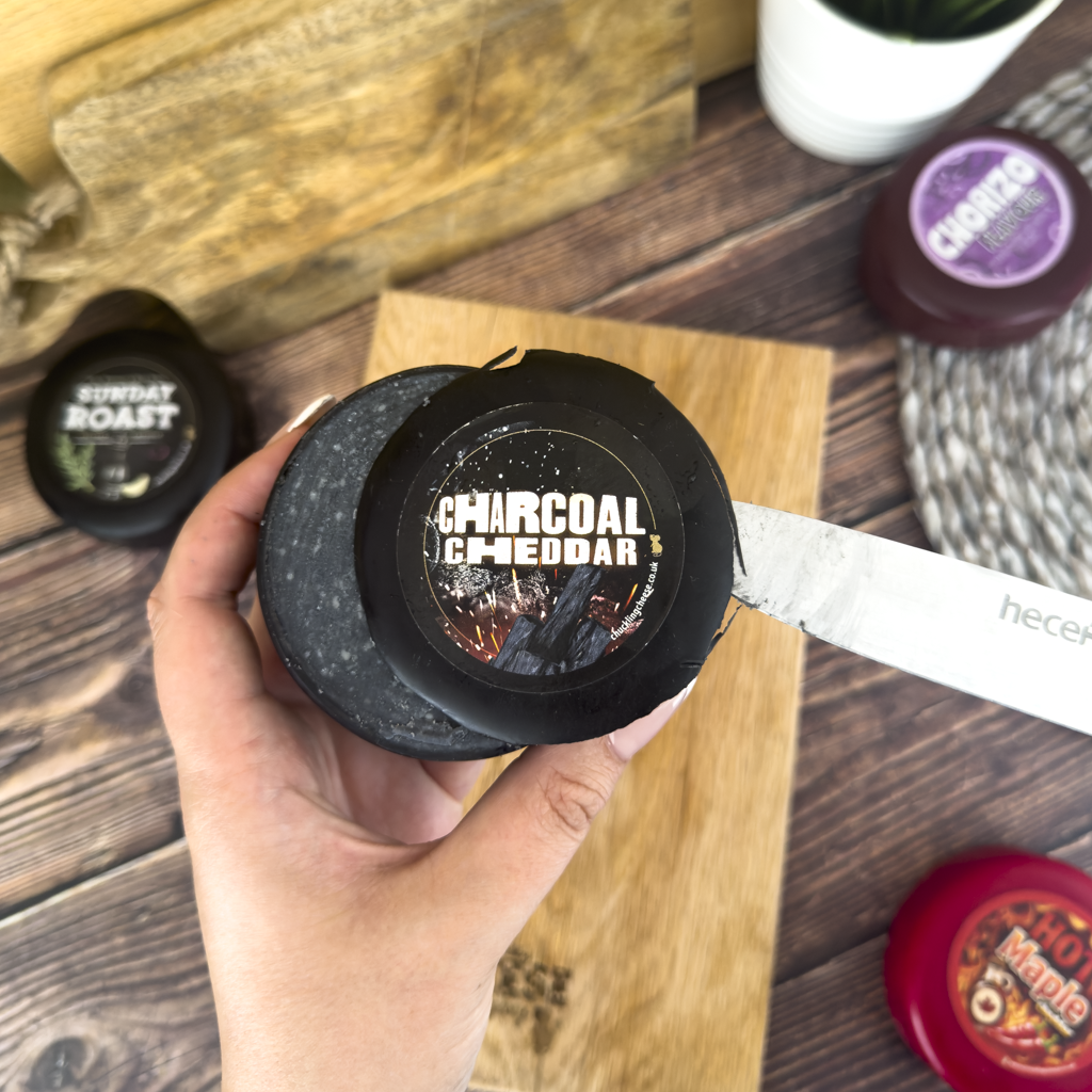 Charcoal Cheddar cheese truckle being sliced with a knife on a wooden board, surrounded by other Chuckling Cheese truckles.