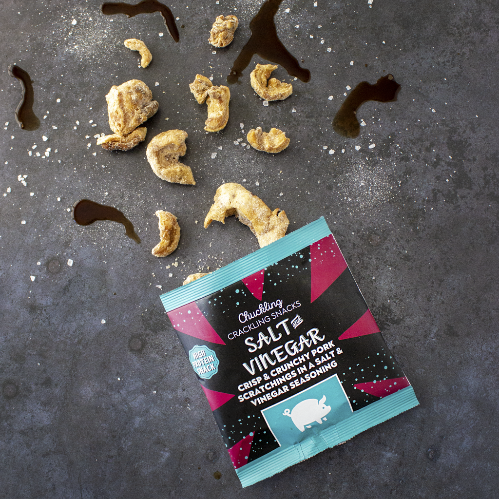 Chuckling Cheese Salt & Vinegar pork scratchings packet with crunchy pieces and seasoning on a dark background