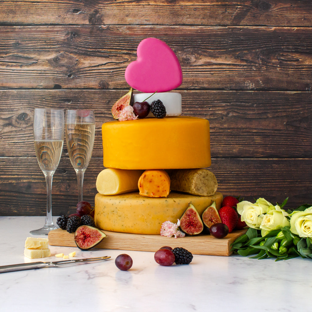 Chuckling Cheese celebration cheese cake tower with stacked cheese wheels, pink heart topper, berries, figs, flowers and champagne glasses