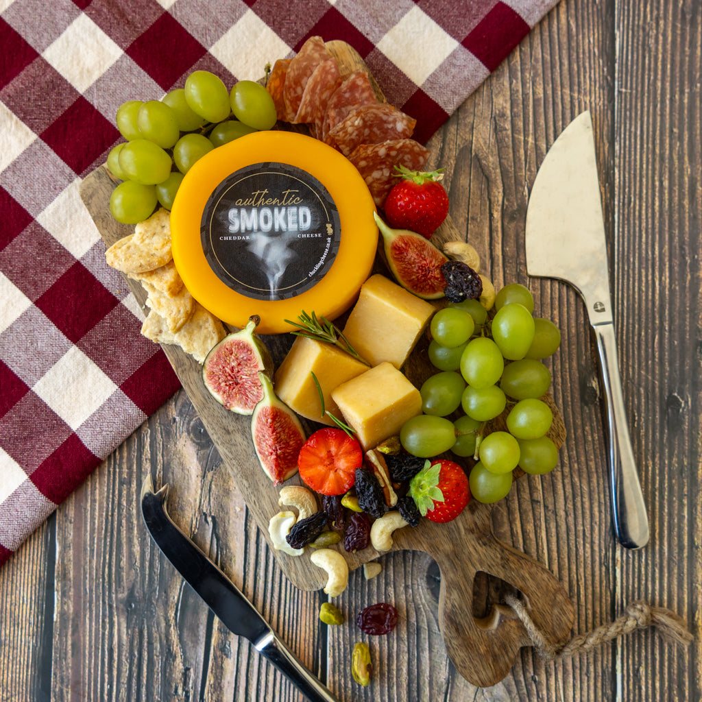 A styled charcuterie board with assorted cheeses, green grapes, figs, strawberries, nuts, crackers, salami, and two knives on a rustic wooden table, with a red-and-white checkered cloth in the background. A round cheese wheel in the center shows the brand name 'Authentic Smoked Cheese'.