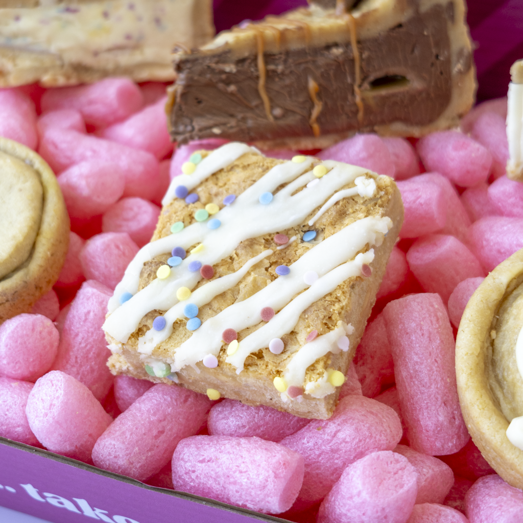 A close-up of assorted gourmet treat box. The central bar is a square, golden-brown blondie slab, topped with white chocolate drizzle and multicolored sprinkles.