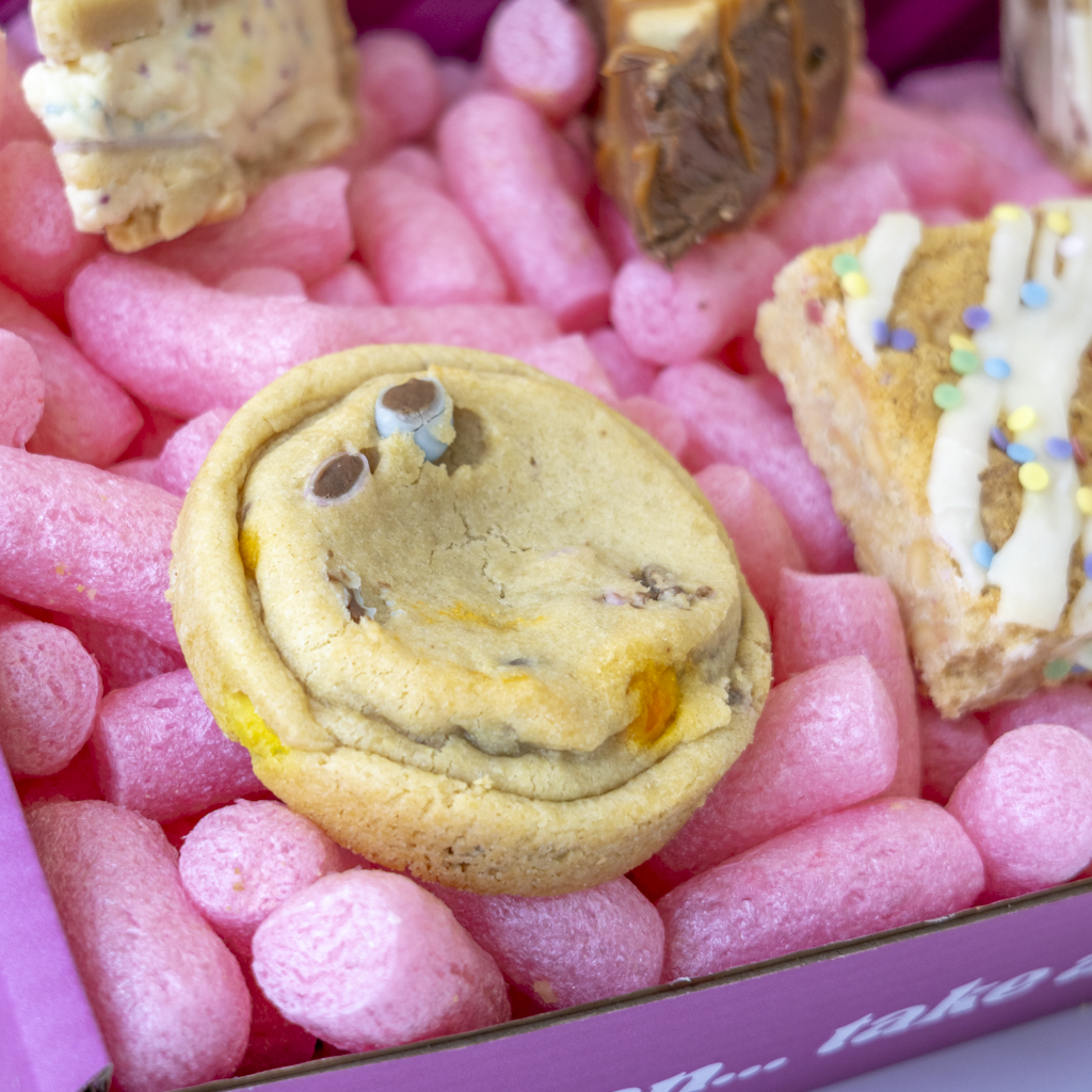A close-up of a pink gift box filled with assorted treats, including a round chocolate bean cookie in the centre and decorated dessert slabs and pie slices at the edges.