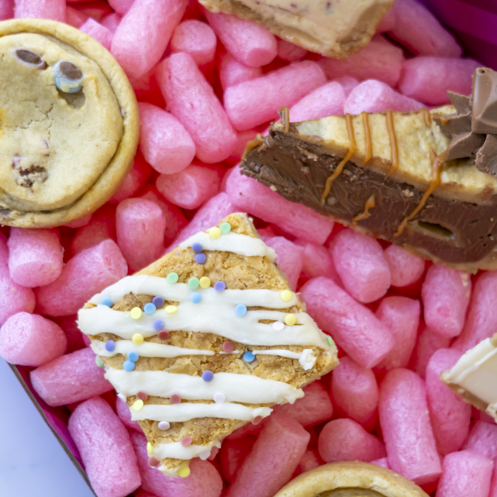 A close-up of assorted cookies and cookie pie slices arranged on pink packing peanuts. Visible pieces include a chocolate bean cookie, a slice of chocolate cake with drizzle, and a square cookie topped with white icing and multicolored sprinkles.