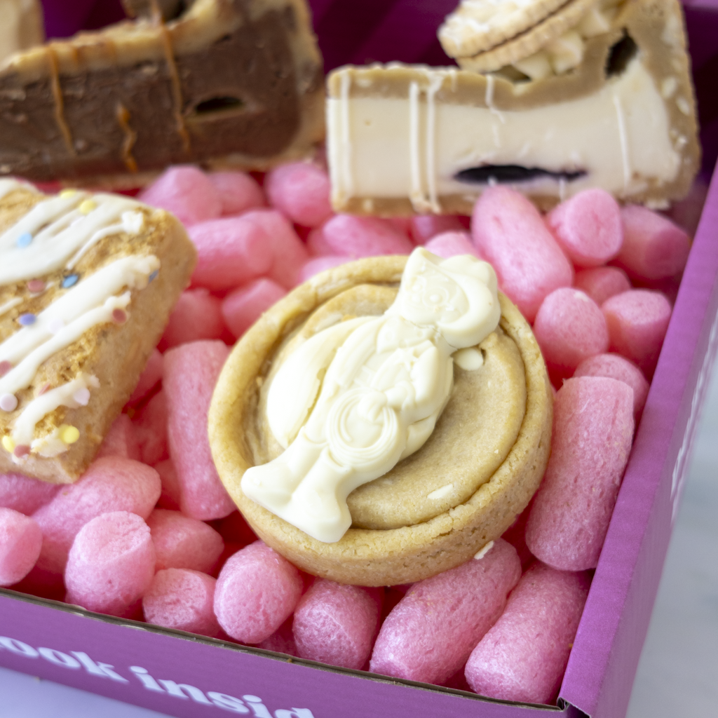 A close-up of a pink cookie box. In the centre is a round golden cookie topped with a Milkybar, surrounded by other assorted sweets and cookies inside a pink box.