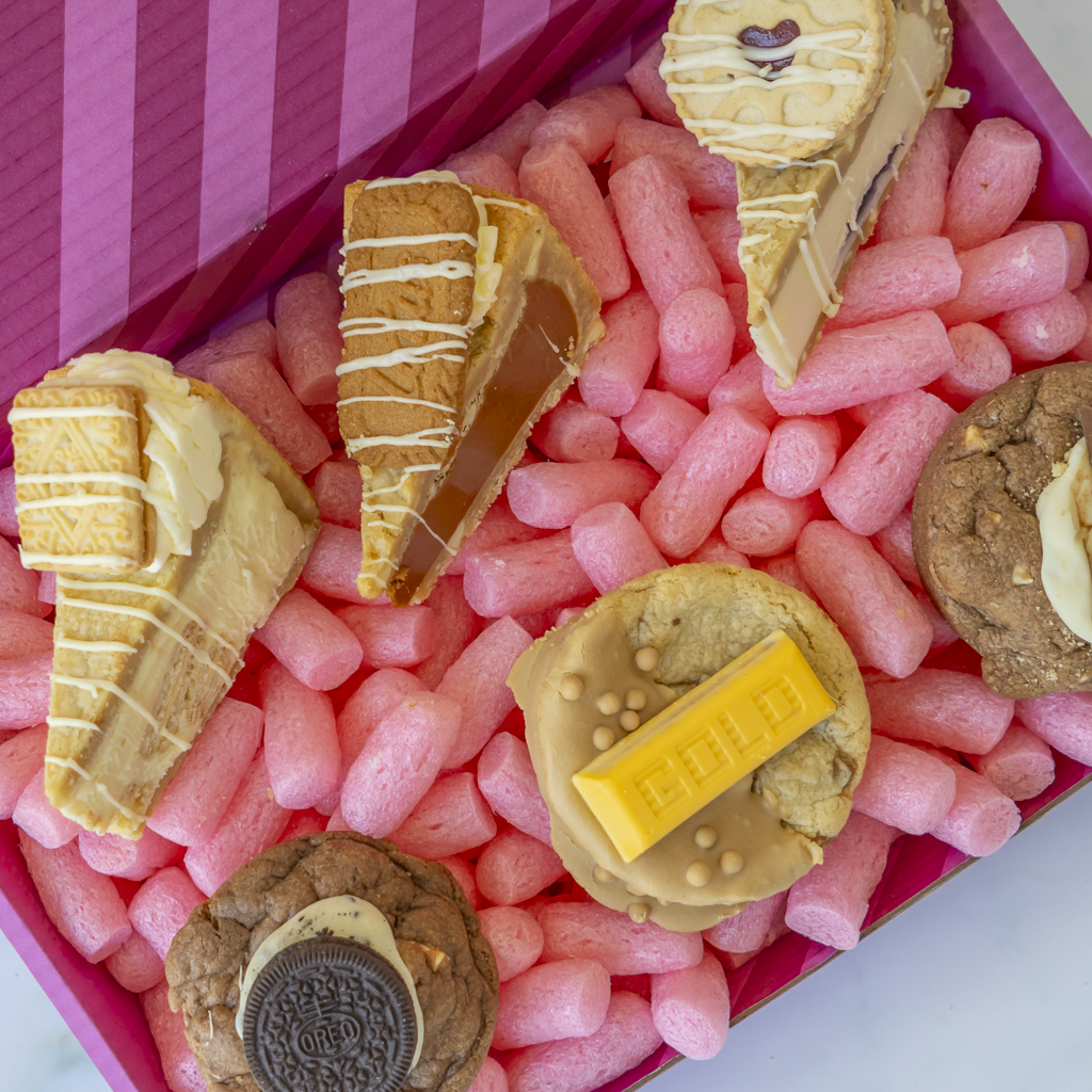 A close-up overhead view of assorted sweet treats arranged in a pink box. The treats include several cookie pie slices with drizzles, cookies, and a chocolate bar, photographed from above inside the box.