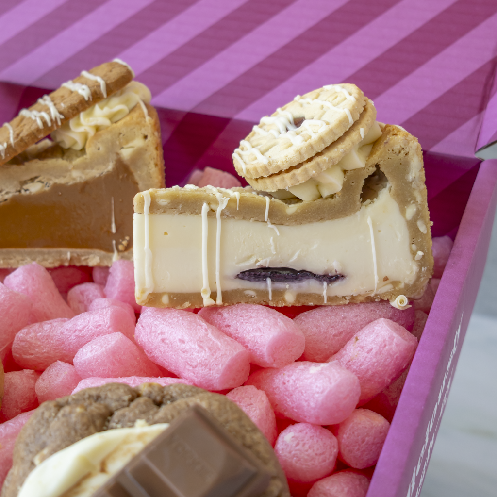 A close-up of assorted gourmet chocolates and cookies presented in a pink box. The center chocolate piece is cut to show a creamy white filling, a dark jam center, and a Jammie Dodger biscuit on top.