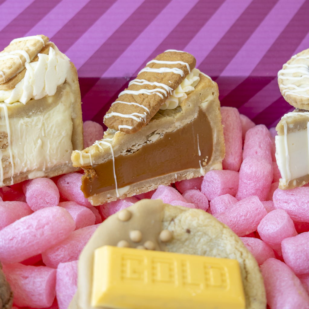 A close-up of assorted gourmet chocolate treats nestled in pink box, including a slice of caramel pie with biscuit topping, custard cream desserts, and a yellow chocolate bar piece.