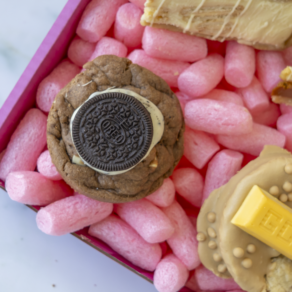 A close-up overhead shot of a cookie gift box containing a chocolate sandwich cookie topped with an Oreo cookie, with other decorated cookies partially visible in a pink box.