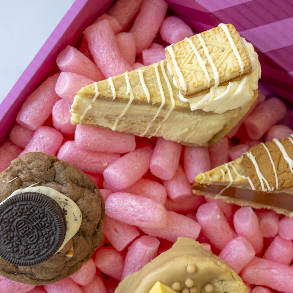 A close-up of assorted baked treats arranged in pink packing peanuts inside a pink box: a cookie pie slice with white drizzle and cookie pieces, a chocolate cookie with cream and Oreo-style filling, and partial views of other layered sweets.