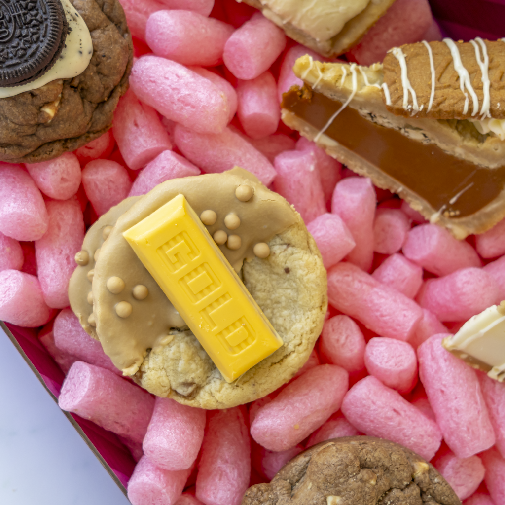 A close-up overhead view of assorted gourmet cookies arranged in a cookie box. The central cookie is topped with a yellow chocolate gold bar, surrounded by other cookies including one with an Oreo and one with biscuit and white drizzle.