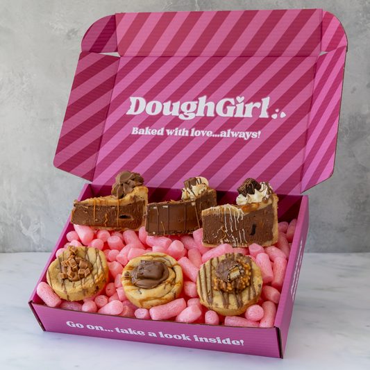 An open pink DoughGirl baking box filled with assorted chocolate-covered cookie pie slices and cookies, on a light marble surface against a gray wall.
