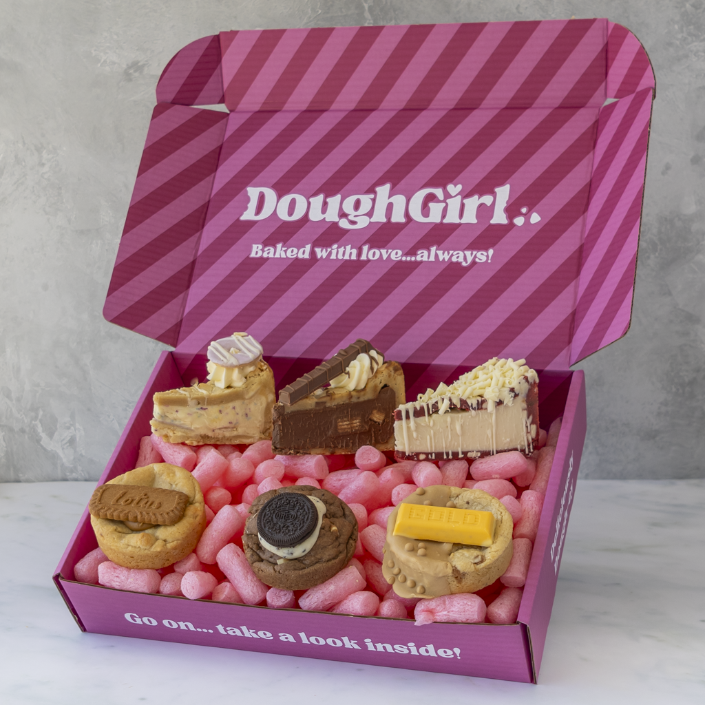 An open pink DoughGirl baking gift box filled with six cookie pie slices and cookie treats, photographed on a light surface against a grey wall.