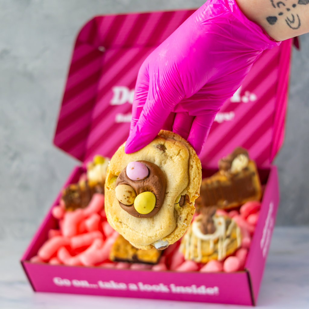 Hand wearing a pink glove holding a Mini Egg Deluxe Cookie with chocolate and colorful Mini Eggs, with a pink box in the background.