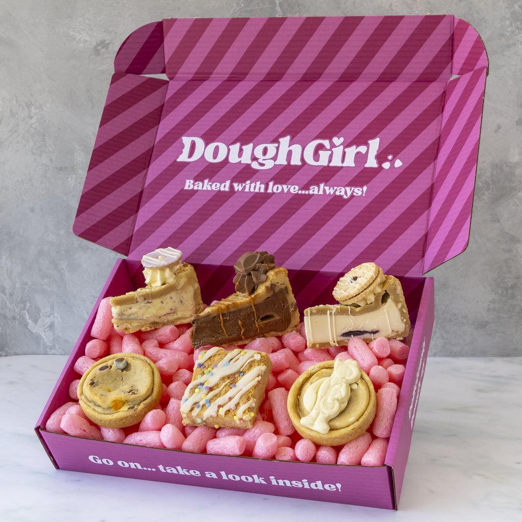 A pink striped DoughGirl baking gift box filled with assorted baked goods, including cookies, cookie pie slices, and a slab, set against a light grey marble surface and textured grey background.