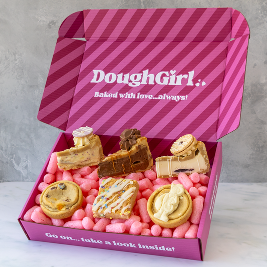A pink striped DoughGirl baking gift box filled with assorted baked goods, including cookies, cookie pie slices, and a slab, set against a light grey marble surface and textured grey background.