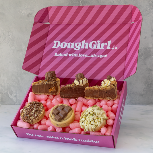 An open pink gift box with diagonal pink stripes and the brand name 'DoughGirl' printed on the inside lid. The box is filled with assorted baked goods, including chocolate cookie pie slices, a chocolate-covered cookie, a nut-topped pastry, and a cream-topped pastry.