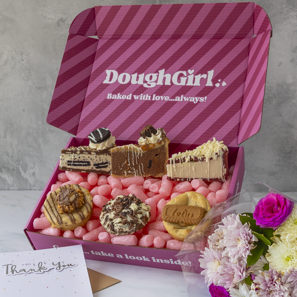An open pink striped gift box branded DoughGirl filled with cookies and cookie pies, surrounded by flowers and a thank-you card.