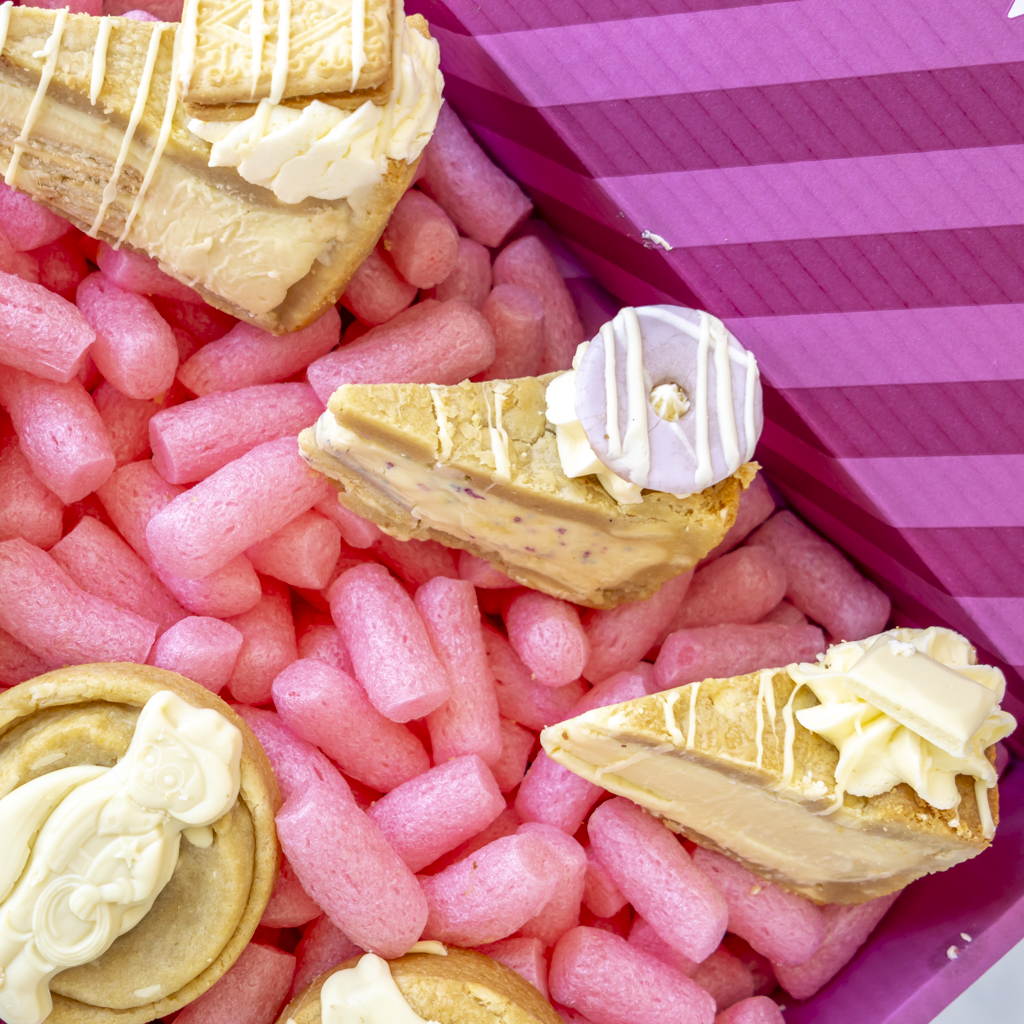 A close-up overhead view of assorted pastries arranged in pink packing foam inside a purple box. Visible items include cream-filled cookie pie slices with drizzle, a slice pastry topped with Party Popper.