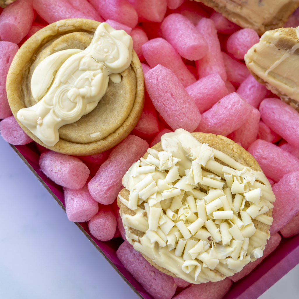 Two decorated cookies in a pink box. One cookie is topped with Milkybar, and the other is topped with shredded white chocolate.