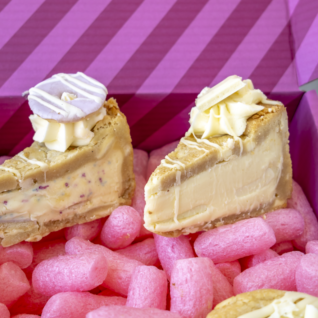 Two decorated dessert slices in a pink gift box. One slice has a light purple glaze and drizzle, and the other has a creamy off-white glaze with drizzle.