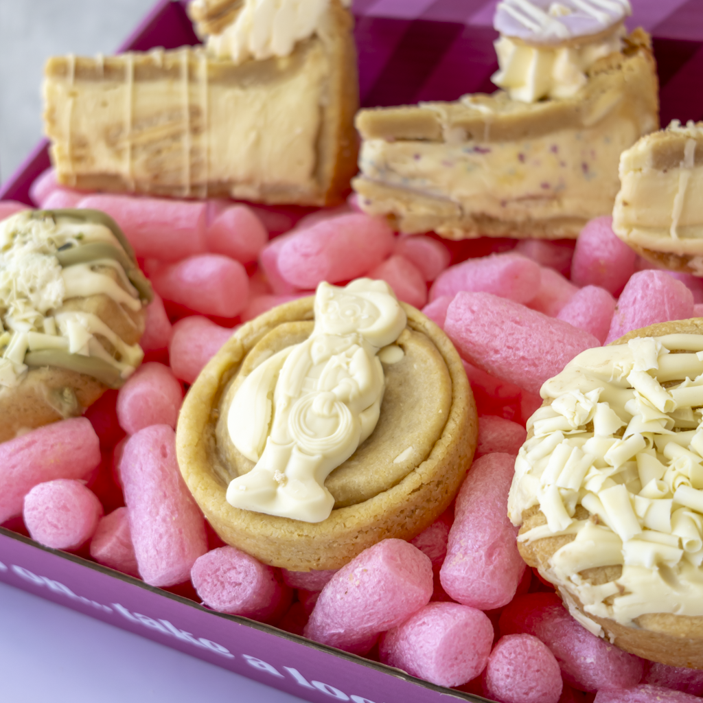 A close-up of assorted baked goods arranged in a pink box. The assortment includes a round chocolate chip cookie with a white chocolate figure, a chocolate-dipped pastry topped with white chocolate shavings, and other chocolate pieces.
