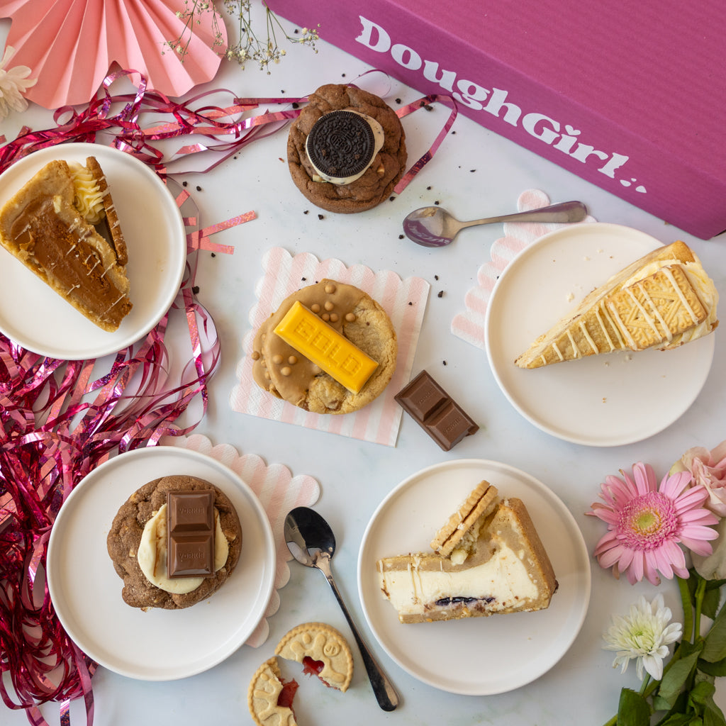 A styled flat-lay of assorted baked goods on white plates, including slices of pie, cookies, and a cheesecake, arranged with pink paper fans, tinsel, flowers, and a pink DoughGirl box in the background.