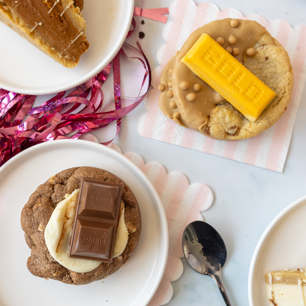 Three chocolate chip cookies decorated with various toppings, including a yellow chocolate bar, a milk chocolate bar, and cookie pie slices, arranged on a white surface with pink and white scalloped paper and a spoon.