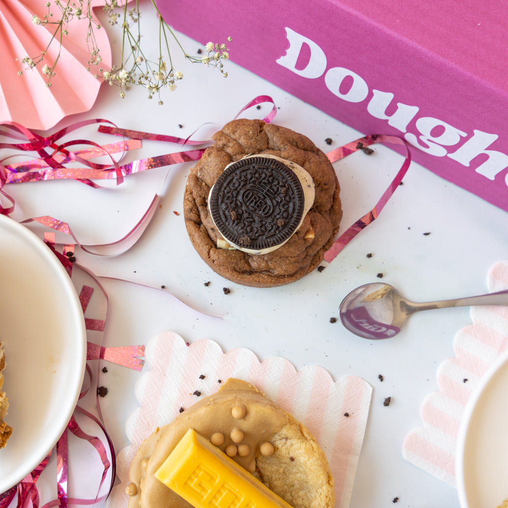 A styled flat-lay of two gourmet cookies on a white surface with pink and white paper fans, ribbons, a spoon, and a pink box labeled 'DoughGirl'. The top cookie is chocolate chip with an Oreo cookie on top, and the bottom cookie is peanut butter chip with a yellow gold bar on top.