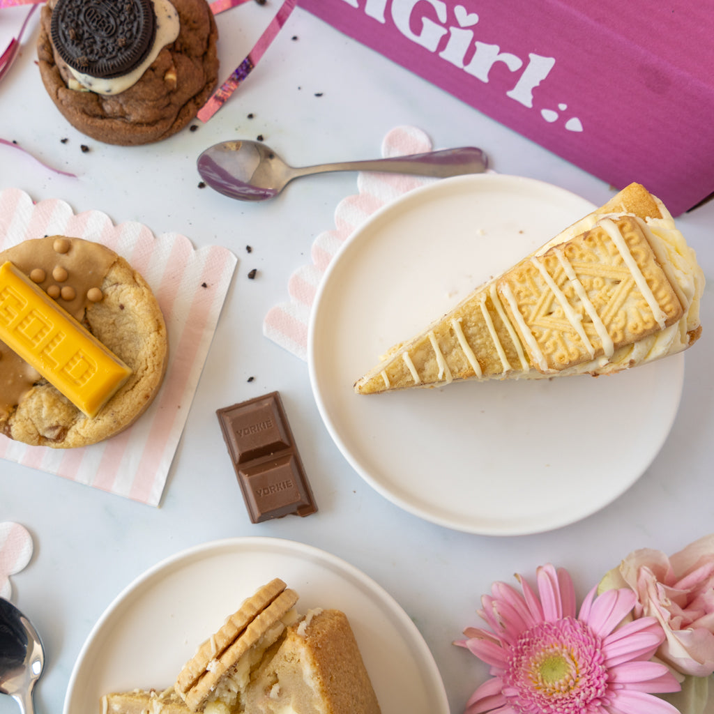 A styled flat-lay of dessert slices on white plates, including a biscuit and cream tart and a chocolate cookie with a biscuit topping, surrounded by pink napkins, a spoon, a chocolate bar, flowers, and a pink branded box.