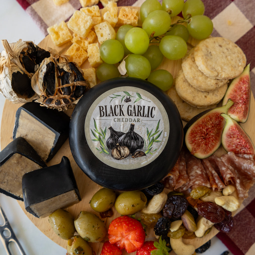 A round wheel of black garlic cheddar cheese sits on a charcuterie board surrounded by green grapes, crackers, figs, black garlic, olives, strawberries, nuts, and cured meat. The cheese label reads 'BLACK GARLIC CHEDDAR'.