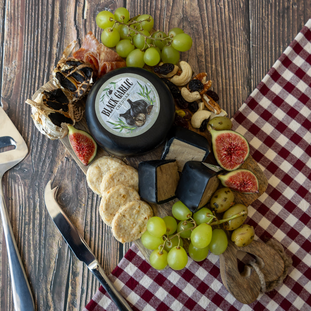 A round wheel of Black Garlic Cheese from San Marcos is styled on a rustic wooden board with crackers, green grapes, figs, nuts, and a red-and-white checkered cloth.