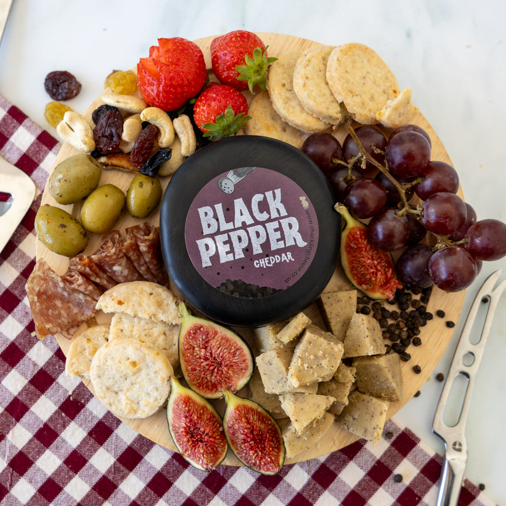 A round wooden charcuterie board arranged with a black cheese wheel labeled 'BLACK PEPPER CHEDDAR' in the center, surrounded by strawberries, grapes, figs, olives, crackers, cheese cubes, and cured meat on a red-and-white checkered cloth.
