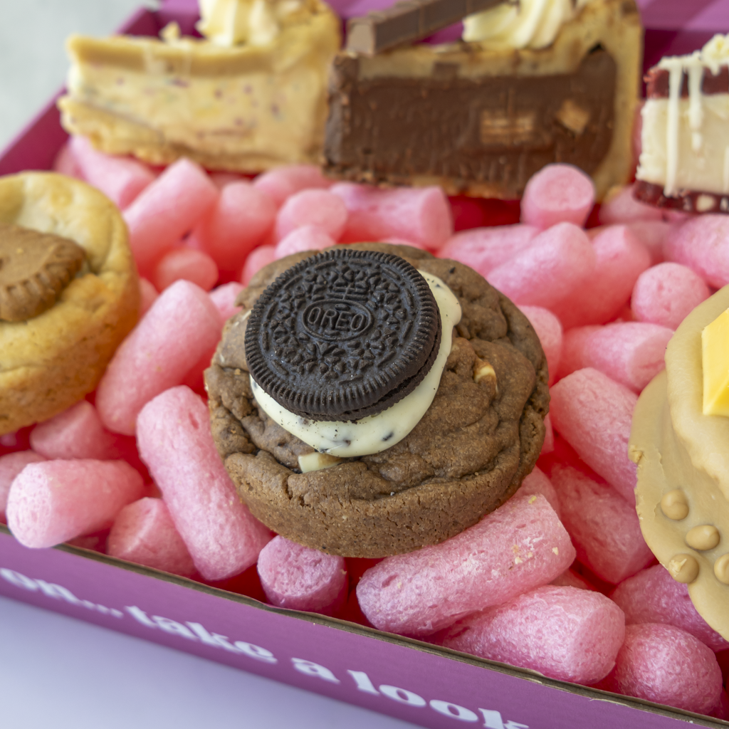 A close-up of a dessert gift box featuring a chocolate cookie topped with a cream-filled filling and an Oreo cookie in the center, with assorted pastries visible in the background.