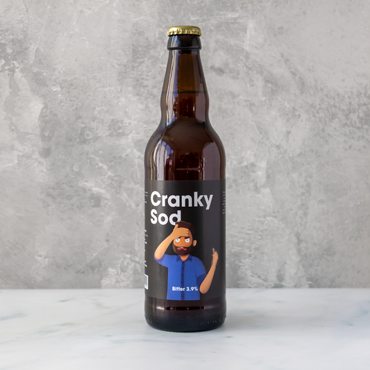 A single brown glass beer bottle with a black label reading 'Cranky Soda' and a cartoon man illustration, standing on a light marble surface against a textured gray wall.