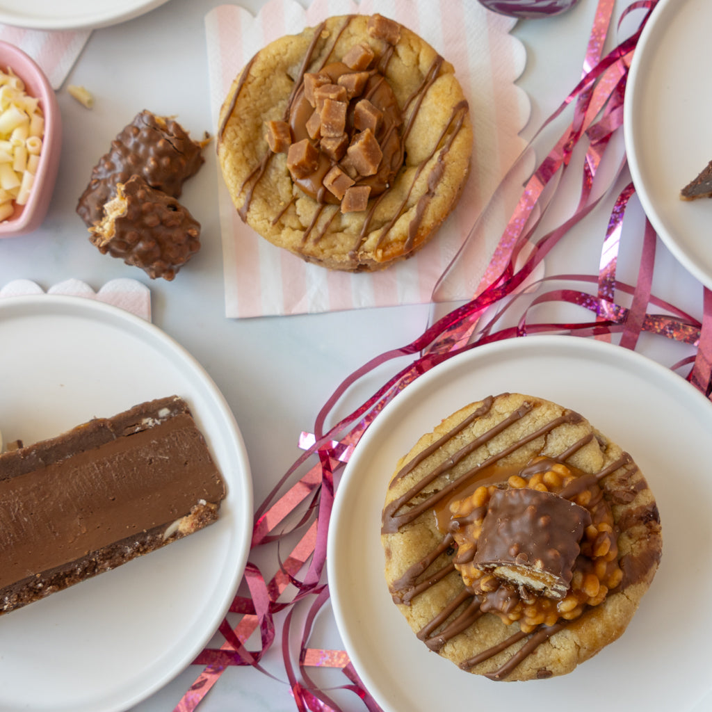 A styled flat-lay of chocolate and caramel cookies on white plates, with pink ribbon streamers and chocolate bar decorations on a light tabletop.