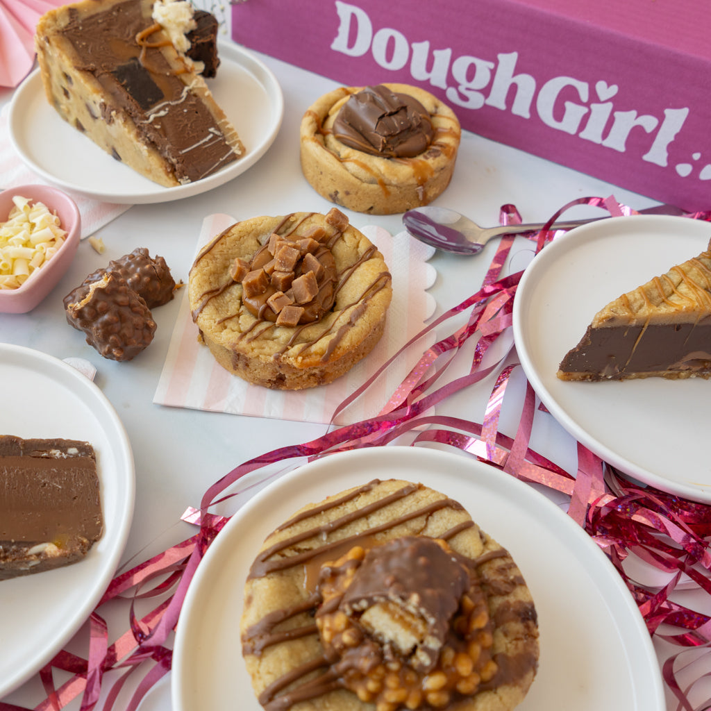 A styled food presentation featuring several chocolate-topped cookies and cookie pie slices arranged on white plates with pink ribbon garlands and a pink box labeled DoughGirl.