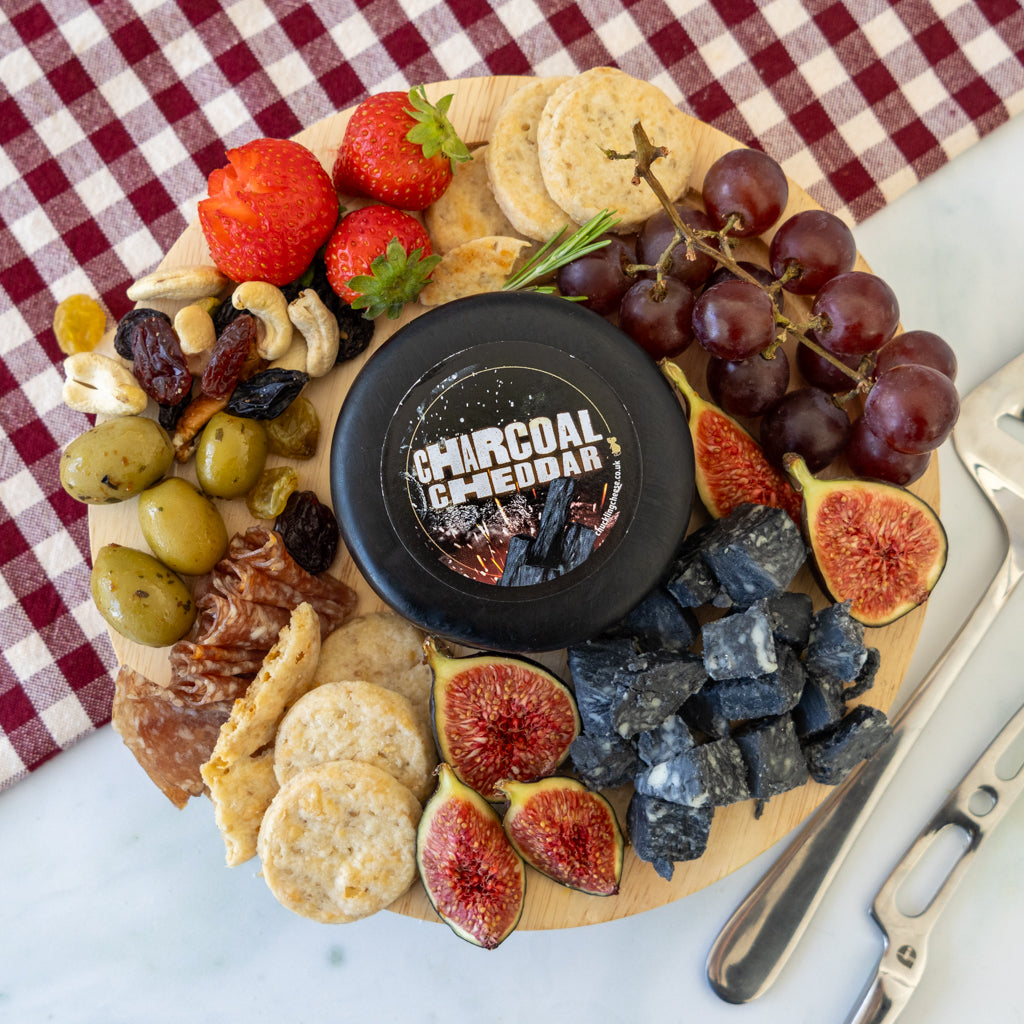 A charcuterie board arranged on a round wooden platter with a black round cheese labeled 'CHARCOAL CHEDDAR,' surrounded by strawberries, grapes, figs, crackers, olives, nuts, dried fruit, and cheese cubes on a white surface with a red-and-white check cloth.