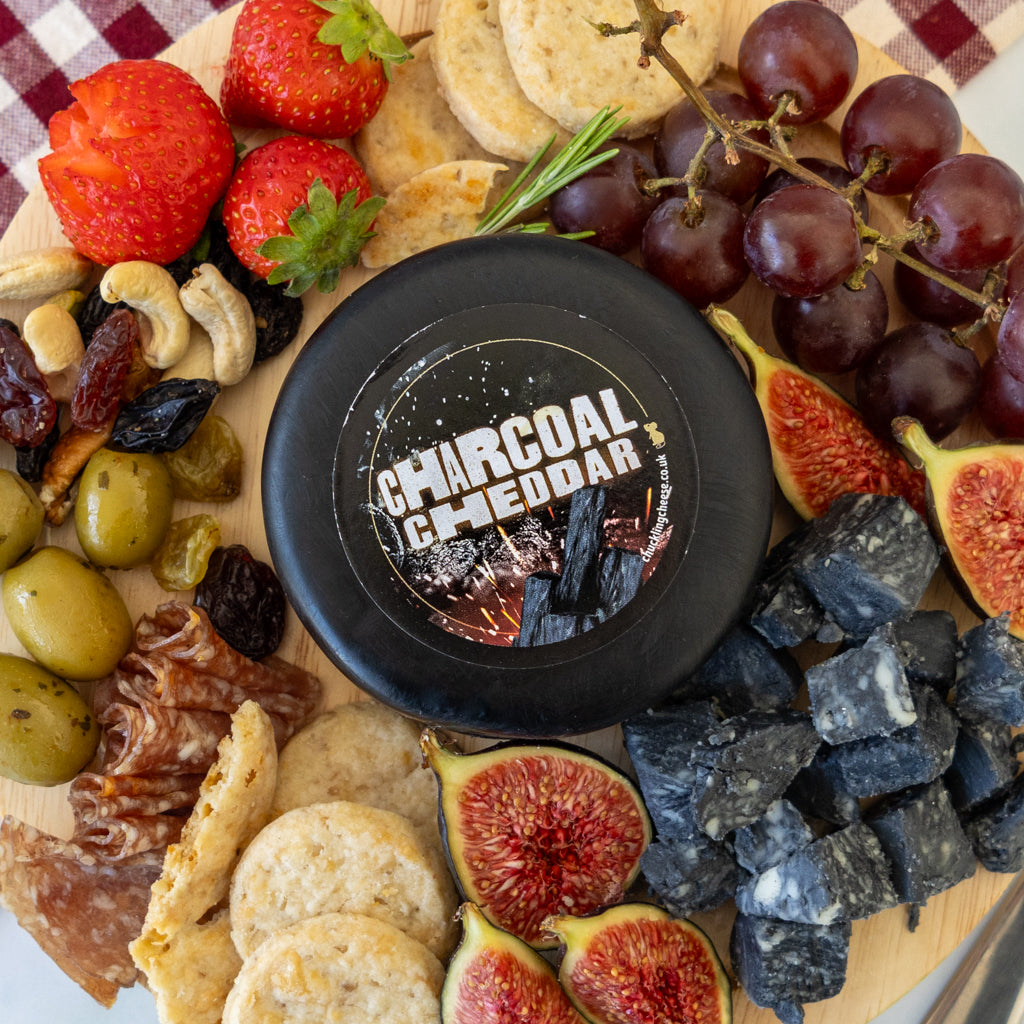 A round black cheese wheel labeled 'CHARCOAL CHEDDAR' arranged on a wooden board with strawberries, grapes, figs, olives, crackers, nuts, and charcoal cubes.