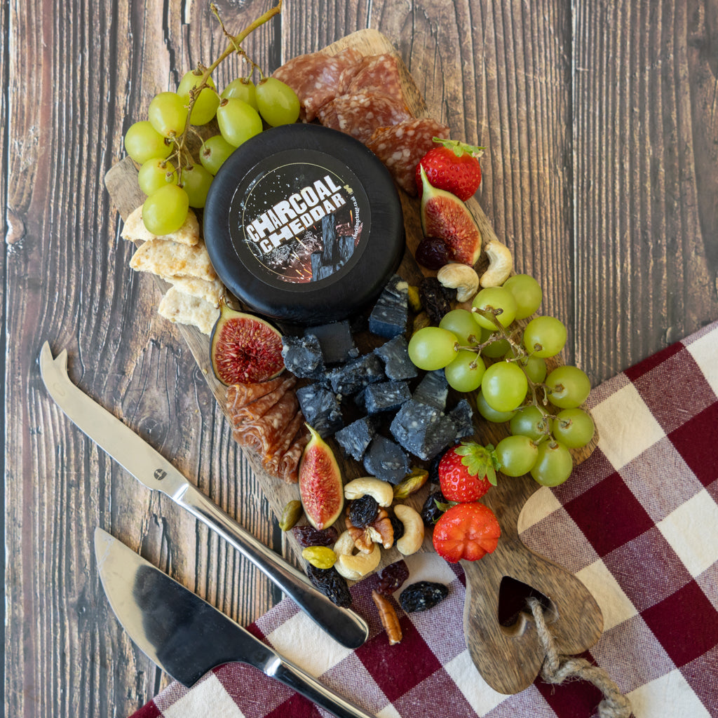 A charcuterie board styled with a round black cheese labeled 'CHARCOAL CHEESE' in the center, surrounded by green grapes, figs, strawberries, nuts, crackers, and cured meats on a wooden board, with a red-and-white checkered napkin and two knives on a rustic wood tabletop.
