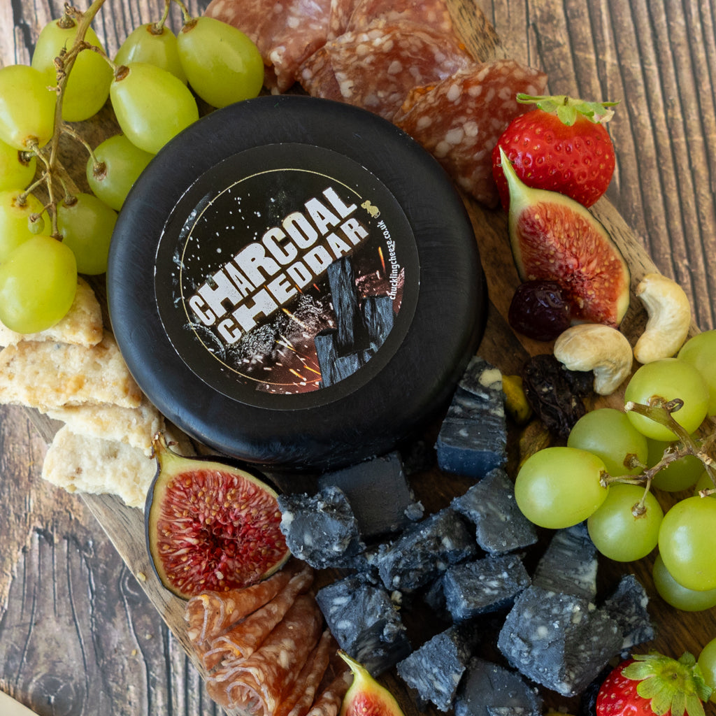A round black wheel of charcoal cheddar cheese is arranged on a wooden charcuterie board with green grapes, sliced figs, strawberries, crackers, salami, and dark cheese cubes. The cheese label reads 'CHARCOAL CHEDDAR'.