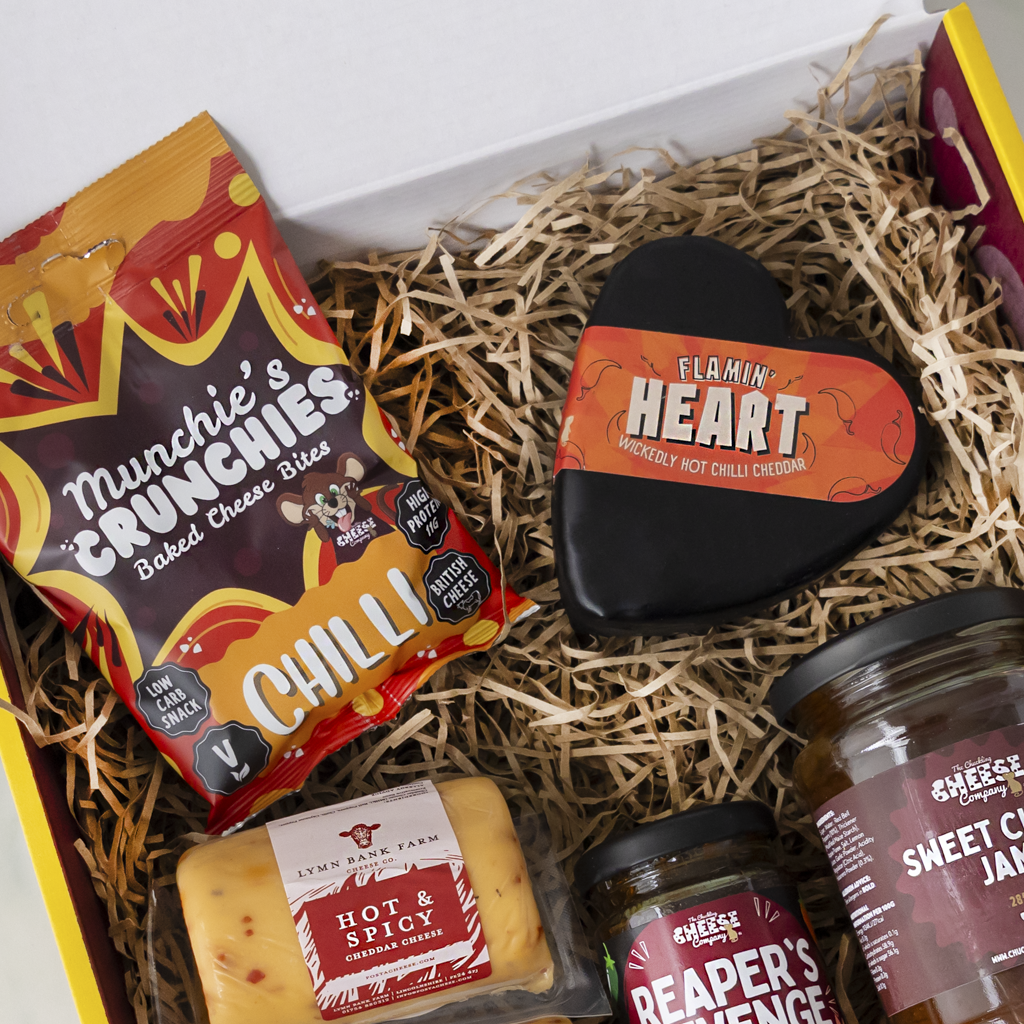 A curated cheese and snack gift box filled with shredded paper, featuring a bag of Munchie's Crunches Chilli baked cheese bites, a heart-shaped Flamin' Heart hot pepper rub, a block of Lyne Star Farm Hot & Spicy cheese, a jar of Sweet Cheddar Jam, and a jar of Reaper's Revenge cheese.