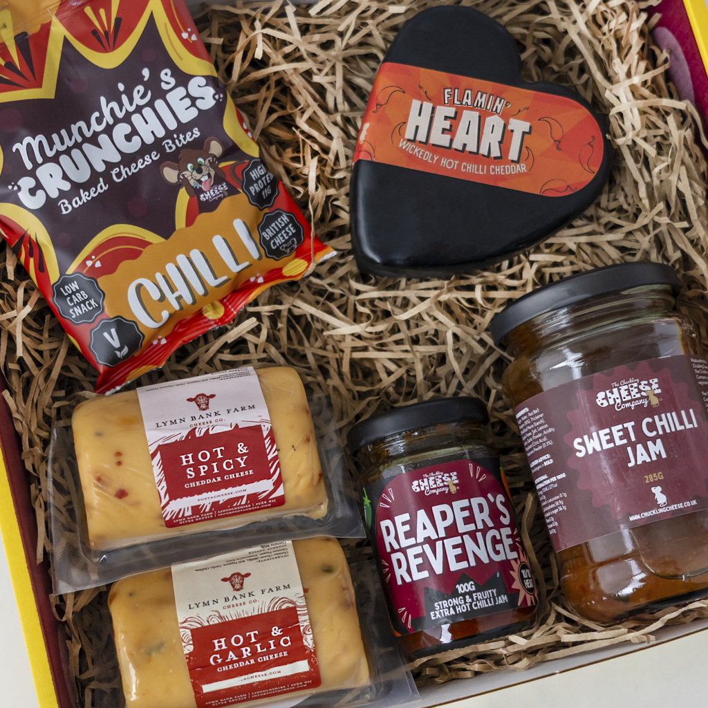 A curated food gift box filled with shredded paper, containing a bag of Munchie's Crunchies baked cheese bites, two packages of Lymn Bank Farm cheese, a heart-shaped hot chilli cheese spread container labelled 'Flammin' Heart', a jar of Reaper's Revenge hot chilli jam, and a jar of Sweet Chilli Jam.