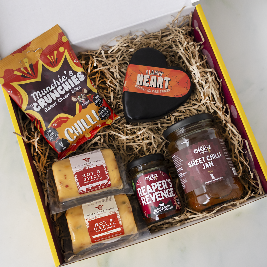 A gourmet gift box filled with cheese and snack items, including Munchie's Crunchies Chilli, a heart-shaped cheese block, Reader's Revenge cheddar, and Sweet Chilli Jam, arranged in shredded paper inside a yellow and white box.