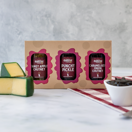 Three small jars of chutney in a cardboard box with a block of cheese and bowl on a table.