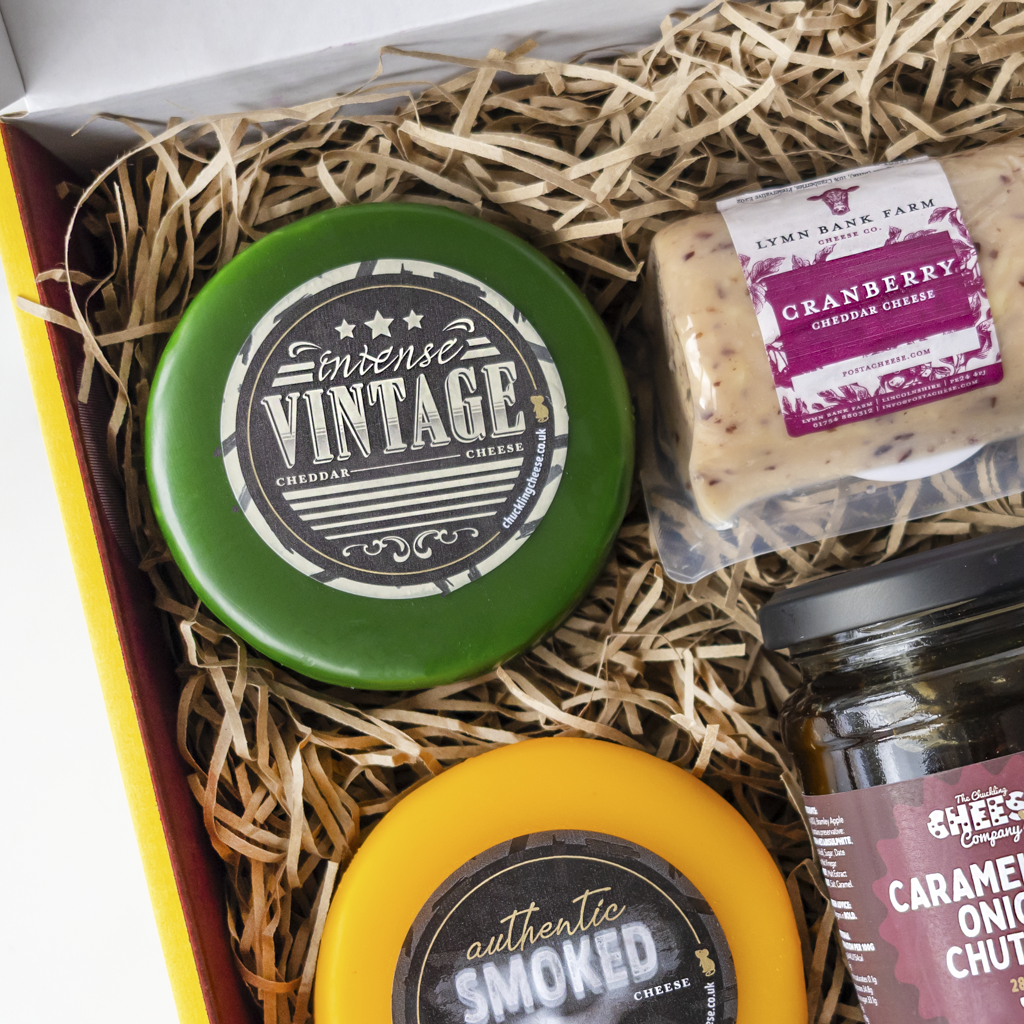 A gourmet cheese gift box filled with shredded paper straw, featuring a green round cheese labeled 'Intense Vintage', a packaged block of 'Cranberry Cheddar Cheese' from Lymn Bank Farm, a jar of caramel onion chutney, and a partially visible yellow smoked cheese round.