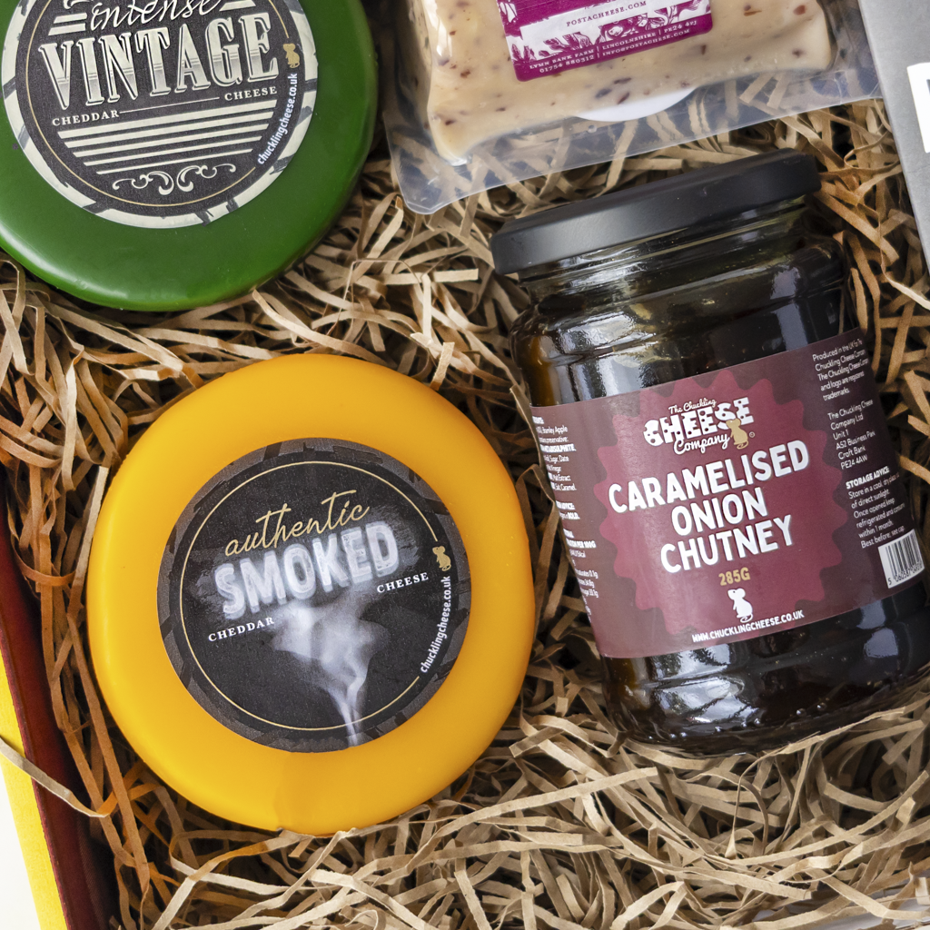A gourmet cheese and chutney gift box featuring a round green wheel of Intense Vintage Cheddar, a yellow wrapped wheel of Authentic Smoked Cheddar, a glass jar of Caramelised Onion Chutney, and a packaged cheese slice nestled in straw packaging.